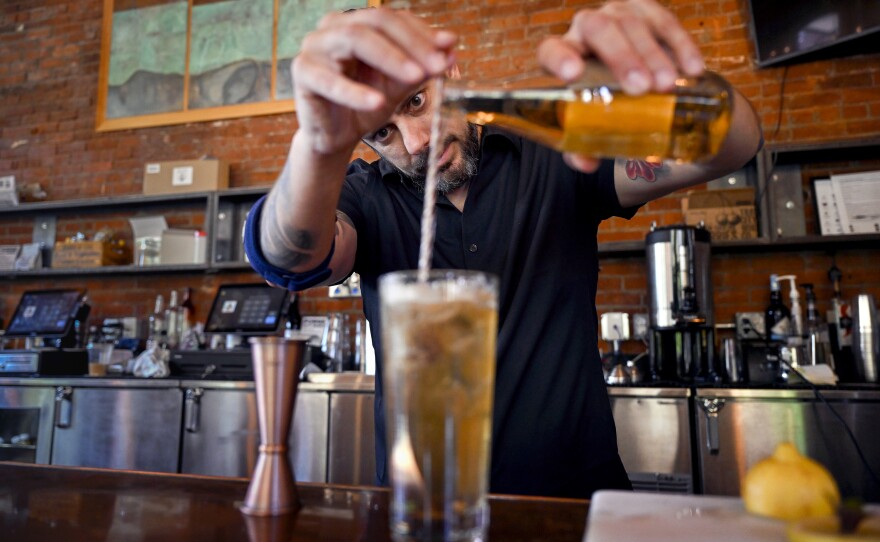 Justin Morales behind the bar, stirring a tall iced cocktail while pouring from a bottle